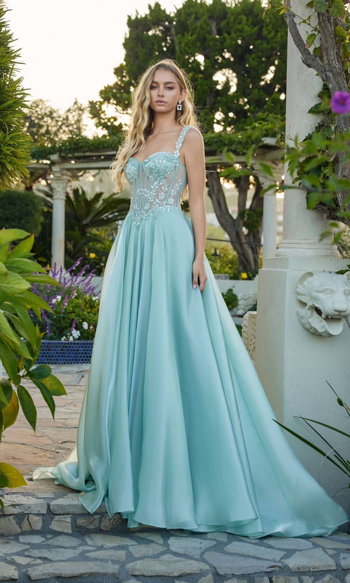 A woman stands outdoors in the sage green Long Prom Dress: Juliet JT2639G, featuring an elegant a-line satin skirt and sheer embellished corset bodice, with lush greenery and stone columns in the background.